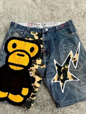 Bape camo shorts med ape-motiv - Unika denimshorts från Bape med camo-detaljer, stor gul och svart ape-figur på vänster ben och vit blixtformad applikation på höger ben. Shorts med streetwear-vibe och tydliga grafiska tryck, perfekta för dig som gillar statement-plagg.