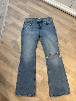 Blå bootcut jeans med slitning - Ett par blå bootcut jeans från Ginatricot med en ripped detalj på höger knä. Jeansen har en ljus blå färg och raka ben med lätt utsvängning. Inga tecken på slitningar eller användning💕
