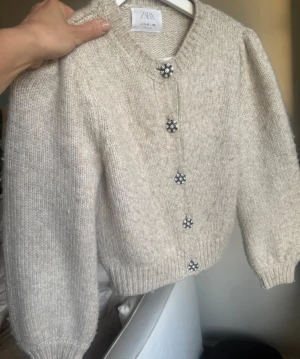 Beige stickad kofta från Zara -  Beige stickad kofta från Zara med puffade ärmar och glittrande blomformade knappar. Koftan har ribbstickade muddar. Lite noppig men annars bra