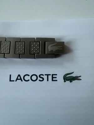  Lacoste armband  - Snyggt armband i silverfärgad metall från Lacoste med klassisk krokodil-logga på en av länkarna. Armbandet har rektangulära länkar med diskret mönster och kommer i original ask. Perfekt accessoar för dig som gillar stilrena detaljer. Den är 20cm men kan enkelt justeras 