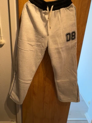 Grå mjukisbyxor med svart midja - Snygga grå mjukisbyxor med svart resårmidja och vitt snöre. Tryck med siffran 08 på ena benet och en bakficka. Sköna och avslappnade med raka ben, perfekta för chill dagar. Materialet känns mjukt och bekvämt.