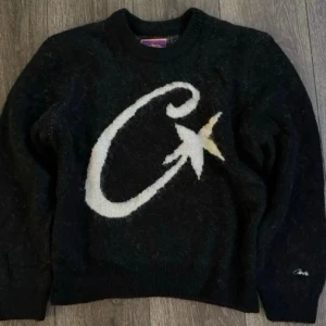 Svart stickad tröja från Carhartt - Stickad svart tröja från Carhartt med stor vit C och stjärna på bröstet. Tröjan har rund halsringning, långa ärmar och ribbade muddar. Materialet ser fluffigt och mjukt ut, perfekt för kyligare dagar. Carhartt-logga broderad på ärmen.