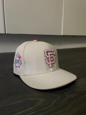 Beige New Era Giants keps med rosa detaljer - Snygg beige keps från New Era med broderad Giants-logga i rosa framtill och rosa detaljer på sidan. Klassisk rak skärm och justerbar passform. Perfekt för dig som vill ha en stilren och sportig look med en twist. Materialet är tålig bomull.