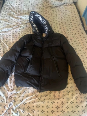 Svart pufferjacka från Moncler - Säljer en svart pufferjacka från Moncler med stor huva där Moncler-loggan är tryckt längs kanten. Jackan har två snedställda dragkedjefickor framtill och är tillverkad i glansigt material. Perfekt för kalla vinterdagar och riktigt snygg streetstil. Storlek passar M och L