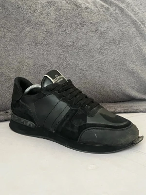 Svarta sneakers från Valentino - Snygga svarta sneakers från Valentino Garavani med klassisk låg profil. Skorna har en mix av läder och mocka, svarta skosnören och coola detaljer på hälen. Perfekt för dig som vill ha en stilren och exklusiv look.