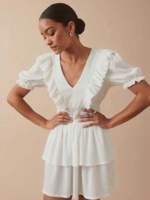 Vit volangklänning med v-ringning - Superfin vit kortklänning med v-ringad hals och volangdetaljer längs axlar och bröst. Klänningen har puffiga kortärmar med rynkade avslut och ett smockat parti i midjan. Materialet är lätt och luftigt, perfekt för varma dagar.