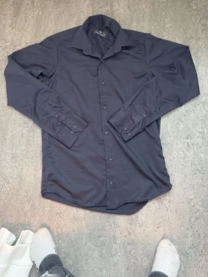 Mörkblå skjorta från YNGmenCLASS - Säljer en stilren mörkblå skjorta från YNGmenCLASS. Skjortan har klassisk krage, knappar hela vägen och långa ärmar med manschettknappar. Perfekt för dig som gillar en enkel och clean look. Materialet känns mjukt och bekvämt.