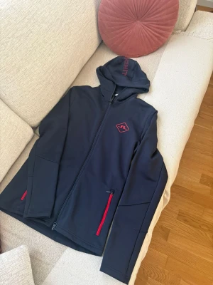 Mörk blå hoodie från J.Lindeberg - Snygg mörk blå hoodie från J.Lindeberg med hel dragkedja och röda detaljer på fickorna. Märkets logga i rött på bröstet och tryck på huvan. Tillverkad i ett mjukt och stretchigt material som passar perfekt till en avslappnad stil.