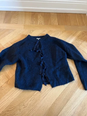 Mörkblå stickad kofta med rosetter - Mysig mörkblå stickad kofta från Gina Tricot med dekorativa rosetter längs öppningen framtill. Koftan har rund halsringning och långa ärmar. Perfekt för lager på lager och en trendig look.