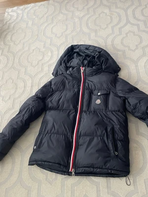 Svart Moncler pufferjacka med huva - Säljer en svart pufferjacka från Moncler.i väldigt bra skick använd bara denna vintern aldrig annars av mig.jackan håller dig väldigt varm och är perfekt för vintern.den är i storlek 4 alltså L skulle säga att den funkar i Xl också.Inget fel på jackan förutom att hängaren är sönder kolla bild 3.den har 2 drag kedjor 1 med franska flagan och 1 bara svart så du man välja mellan de.