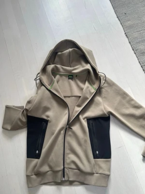 Hugo Boss hoodie - Snygg beige hoodie från Hugo Boss med svarta paneler på sidorna och dragkedjeförsedda fickor. Huva med dragsko och diskret grön detalj på insidan av kragen. Tillverkad i mjukt sweatshirtmaterial och har ribbade muddar vid ärmar och nederkant.