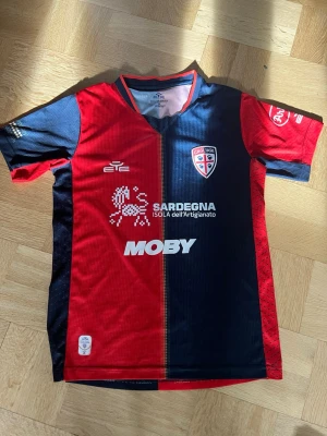 Cagliari Calcio matchtröja röd/blå - Säljer en officiell Cagliari Calcio matchtröja från EYE Sport. Tröjan är kortärmad och har en cool design med röd och mörkblå färgblock, klubbmärke på bröstet och flera sponsorer tryckta på både fram- och baksidan. Materialet är lätt och andas, perfekt för fotboll.