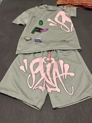 Grått set med rosa graffiti från syna world  - Säljer ett grått tvådelat set från SVRC med coola rosa graffiti-tryck på både tröja och shorts. Setet har flera färgglada patches på tröjan och snörning i midjan på shortsen. Materialet är mjukt och stretchigt, perfekt för en avslappnad streetwear-look.