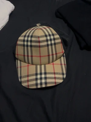 Burberry beige rutmönstrad keps - Snygg keps från Burberry med klassiskt beige rutmönster i svart, vitt och rött. Kepsen har svart mesh baktill och justerbar snapback-spänne för perfekt passform. Perfekt accessoar för dig som vill ha en stilren och trendig look.