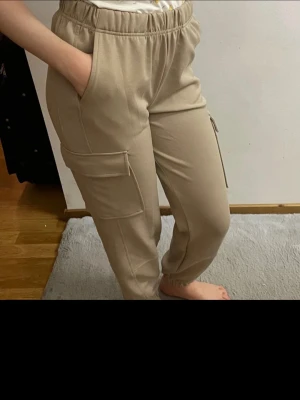 Beiga cargobyxor med resårmidja - Snygga beiga cargobyxor med resår i midjan och vid bensluten. Byxorna har stora fickor på sidorna och är tillverkade i ett mjukt och bekvämt tyg. Perfekta för en avslappnad och trendig look.