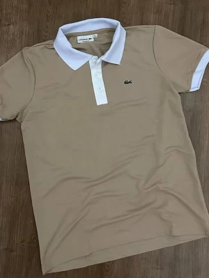 Beige pikétröja från Lacoste - Snygg beige pikétröja från Lacoste med klassisk vit krage och knappslå. Tröjan har korta ärmar och den ikoniska gröna krokodilloggan broderad på bröstet. Perfekt för en stilren och sportig look.