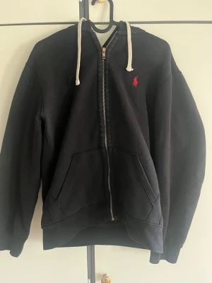 Ralph Lauren zip hoodie - Klassisk svart hoodie från Ralph Lauren med dragkedja framtill, vit dragsko i huvan och den ikoniska röda logotypen broderad på bröstet. Hoodien har känguruficka och ribbade muddar vid ärmar och nederkant. Perfekt för en avslappnad och stilren look.