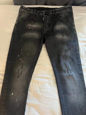 Svarta jeans med färgstänk och slitningar - Snygga svarta jeans med coola slitningar och färgstänk i vitt och rött på benen. Jeansen har klassisk femficksmodell och rak passform. Perfekta för dig som gillar en trendig och avslappnad streetstil.