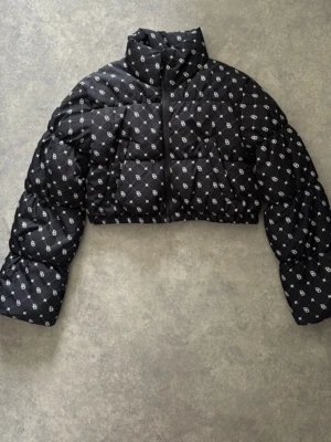Svart cropped pufferjacka med mönster - Svart cropped pufferjacka från SikSilk med heltryckt vitt logomönster. Jackan har hög krage, dragkedja framtill och quiltad design. Perfekt för att hålla värmen och sticka ut i vinter. Materialet är glansigt och ger en modern look.