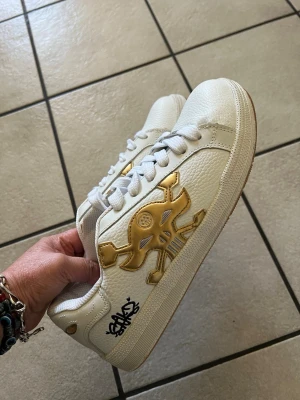 Vita sneakers med gulddetaljer - Unika vita sneakers från Philipp Plein med stora guldfärgade dödskalle-detaljer på sidan och hälen. Skorna har vita snören, svart logga på sidan och är tillverkade i läder med gummisula. Perfekta för dig som vill sticka ut med en edgy och lyxig stil.