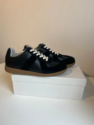 Svarta Maison Margiela sneakers - Maison Margiela sneakers i svart skinn och mocka med kontrasterande vita snören och beige gummisula. Klassisk rund tå och platt sula, stilren design med diskreta paneler och mjuk insida. Perfekt för dig som gillar minimalistisk och clean look. Hör av er vid någon fråga eller fundering. Priset kan diskuteras vid snabb affär!✅