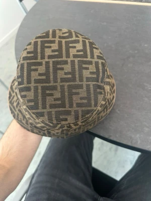 Fendi bucket hat med FF-mönster - Säljer en snygg bucket hat från Fendi i brunt tyg med det klassiska FF-logomönstret över hela hatten. Hatten har en mjuk form och är tillverkad i ett slitstarkt tygmaterial. Perfekt för dig som vill sticka ut med en ikonisk accessoar.