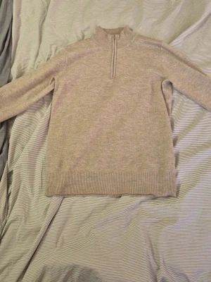 Beige stickad tröja från RYVLS - Stickad tröja i beige från RYVLS Stockholm med halv dragkedja framtill och ribbade muddar vid ärmslut och nederkant. Tröjan har en avslappnad passform och är perfekt för lager på lager. Klassisk och enkel design som passar till det mesta.