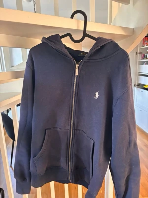 Marinblå hoodie från Polo Ralph Lauren - Snygg marinblå hoodie från Polo Ralph Lauren med dragkedja framtill och klassisk vit logga på bröstet. Hoodien har huva, kängurufickor och ribbade muddar. Tillverkad i mjukt bomullsmaterial som känns skönt mot huden. Den är lite solblekt på framsidan däravpriset