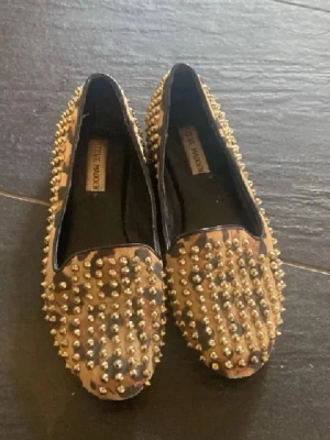 Leopardmönstrade ballerinaskor Steve Madden - Coola ballerinaskor från Steve Madden med leopardmönster och massor av guldfärgade nitar. Skorna har en klassisk rundad tå och är tillverkade i textil med svarta detaljer på insidan. Perfekta för dig som vill sticka ut med din stil.