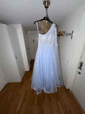 Ljusblå asymmetrisk balklänning - Magisk ljusblå balklänning med ett axelband och transparent tyll över ena armen. Klänningen har vackra broderade blommor och pärlor på livet samt ett svepande, voluminöst kjolparti i flera lager tyll. Perfekt för bal eller speciella tillfällen.