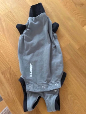 Cat body suit Buster - Grå hundoverall från Buster med svarta detaljer och tryckknappar framtill. Overallen har öppningar för ben och är tillverkad i ett slitstarkt tyg, perfekt för promenader i alla väder. Passar mindre till medelstora hundar.
