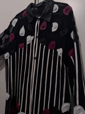 Svart skjorta med ränder och 3D-blommor - Unik svart skjorta med vita vertikala ränder och dekorativa 3D-blommor i vitt, svart och vinrött. Skjortan har klassisk krage, knäppning framtill och lång ärm. Materialet är tunt och transparent, vilket ger en cool och edgy vibe.