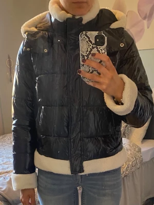 Svart pufferjacka med fuskpälsdetaljer - Säljer en svart glansig pufferjacka med vita fuskpälsdetaljer på krage, ärmslut och nederkant. Jackan har huva och dragkedja framtill. Perfekt för kalla vinterdagar och riktigt mysig att bära. Passar dig som vill ha en varm och trendig vinterjacka. Storlek M men känd som S.