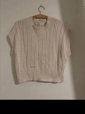 Beige stickad väst med ribbad kant - Säljer en beige stickad väst med bred ribbad kant nedtill och vid halsen. Västen har ett diskret hålmönster och är ärmlös med rund halsringning. Perfekt att styla över en skjorta eller t-shirt! Oanvänd utan prislapp