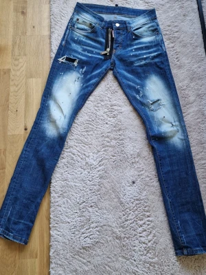 Dsquared2 Tidy Biker Jeans blå distressed strl 42 - Säljer ett par Dsquared2 Tidy Biker Jeans i blå distressed tvätt, storlek 42. Motsvarar ungefär W29 i tum, men kontrollera gärna mått om du vill vara helt säker. Made in Italy. Tvätten har slitningar, blekta partier och färgstänk samt dragkedjedetalj framtill. Se bilder för skick och detaljer.