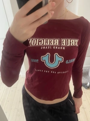 Bordeaux långärmad topp True Religion - Snygg bordeaux långärmad topp från True Religion med tryck framtill i vitt och ljusblått. Modellen är croppad och har en rund halsringning. Perfekt för dig som gillar streetwear och vill sticka ut med ett ikoniskt märke.