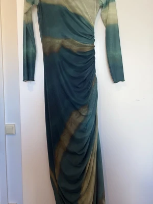 Lång mönstrad klänning med drapering - Snygg långklänning med unikt mönster i blått och beige toner. Klänningen har lång ärm och draperad effekt längs sidan för en cool siluett. Perfekt för dig som vill sticka ut med en trendig look.