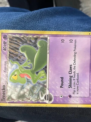 Treecko Pokémonkort holografiskt - Treecko holografiskt Pokémonkort med lila och silver bakgrund. Kortet har 40 HP och visar Treecko i grön färg med röda detaljer. Text och attacker på engelska. Klassisk baksida med Pokéball och blå ram.