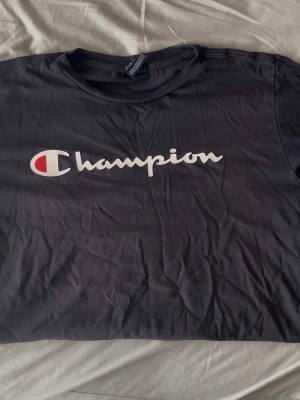 Svart Champion t-shirt med logga - Snygg svart t-shirt från Champion med stor vit och röd logga tryckt över bröstet. Klassisk rund halsringning och korta ärmar. Tillverkad i mjuk bomull som känns skön mot huden. Perfekt för en avslappnad streetwear-look.