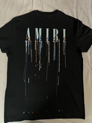 Svart AMIRI t-shirt med färgstänk - Säljer en svart t-shirt från AMIRI med tryck både fram och bak. Stora AMIRI-bokstäver på ryggen och färgstänk i blått, vitt, orange och brunt som rinner ner över tyget. Klassisk passform och rund hals. Perfekt för dig som gillar streetwear och unika detaljer.