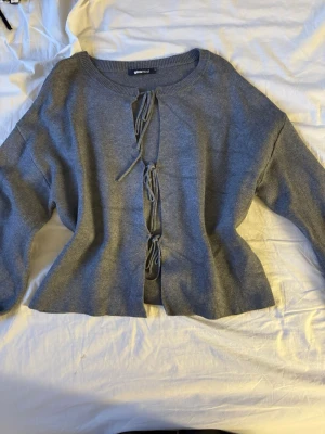 Grå stickad cardigan från Gina Tricot - Säljer en grå stickad cardigan från Gina Tricot med tre knytband framtill. Tröjan har rund halsringning, långa ärmar och är gjord i mjukt stickat material. Perfekt att slänga över en topp för en chill look.