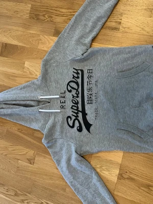 Grå hoodie från Superdry - Snygg grå hoodie från Superdry med stor svart logga och japansk text på bröstet. Hoodien har huva med vita dragsnören och en klassisk magficka. Tillverkad i mjukt bomullsmaterial, perfekt för en avslappnad streetwear-look.