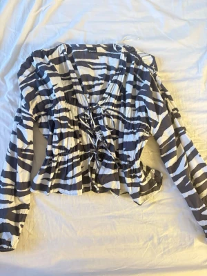Zebramönstrad blus från Gina Tricot - Snygg zebramönstrad blus från Gina Tricot i svart och vitt. Blusen har långa ärmar, v-ringning och knytning framtill. Tillverkad i ett lätt och luftigt material som ger en avslappnad men trendig look.