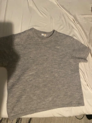 Grå stickad tröja från Gina Tricot - Mysig grå stickad tröja från Gina Tricot med rund halsringning och kortare ärmar. Tröjan har en enkel och stilren design, perfekt att matcha med jeans eller kjol. Materialet är mjukt och ger en avslappnad känsla.