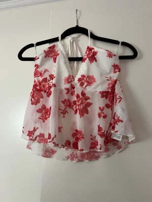 Vit blommig halterneck linnetopp - Söt vit linnetopp med röda blommor och halterneck-knytning i nacken. Toppen har v-ringning och är ärmlös med lätt utsvängd passform. Perfekt för varma dagar och somriga outfits.