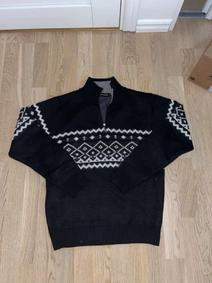 Svart stickad tröja med mönster - Svart stickad tröja med vitt nordiskt mönster över bröstet och ärmarna. Tröjan har lång ärm, ribbade muddar och en half zip-dragkedja vid halsen. Perfekt för kyliga dagar.