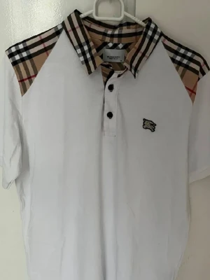 Vit Burberry piké med rutiga detaljer - Vit pikétröja från Burberry med klassiskt rutigt mönster på krage och axlar. Tröjan har korta ärmar, knappar framtill och en liten broderad logga på bröstet. Perfekt för en stilren och trendig look.