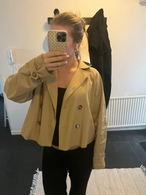 Kort beige trenchcoat med knappar - Snygg kort trenchcoat i beige med dubbelknäppning framtill och bred krage. Jackan har långa ärmar med knappdetaljer vid ärmslut och en avslappnad passform. Perfekt för dig som vill ha en trendig och stilren look.