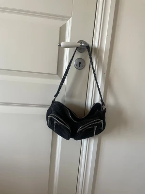 Svart axelväska från Noella - Cool svart axelväska från Noella med flera silvriga dragkedjor och flätat axelband. Väskan har två framfickor med dragkedja och ett stort huvudfack. Perfekt för dig som gillar en edgy och praktisk look.