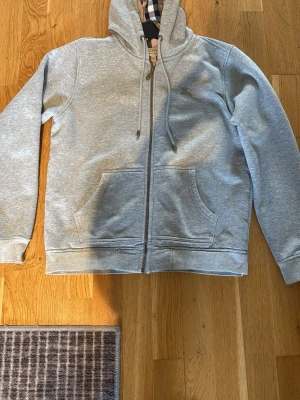 Grå zip-hoodie från Burberry - Snygg grå hoodie med dragkedja från Burberry. Klassisk huva med dragsko och ikoniskt rutigt foder. Mjuk bomullsblandning, två stora fickor framtill och diskret logga på bröstet. Perfekt för en chill och stilren look.